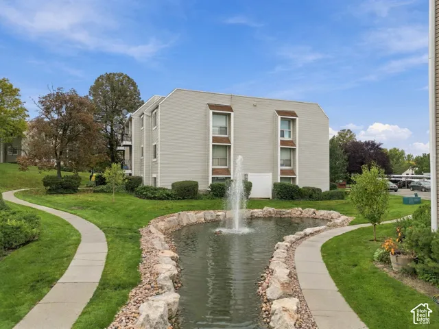 Exterior of multi-story building featuring light-toned horizontal siding, a central water feature with stone-lined perimeter, and paved walkways throughout the landscaped grounds