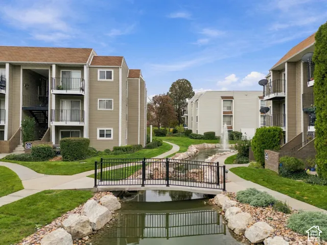 Multi-story residential complex featuring beige siding, private balconies, a central water feature with a stone-lined pond, and a pedestrian footbridge