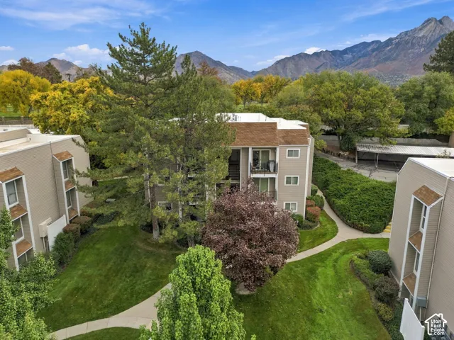 Multi-story building featuring tan siding, a shingled roof, mountain backdrop, mature trees, and manicured landscaping
