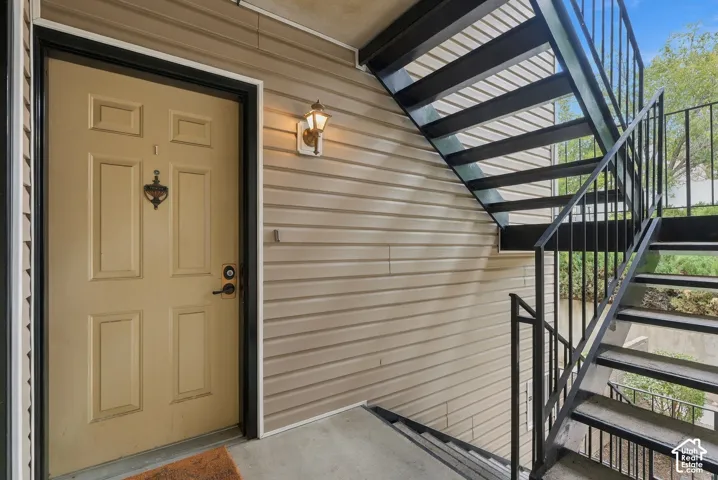 Exterior entryway featuring horizontal siding, a solid-core door with a metal knocker, and an attached wall-mounted lantern light fixture, accented by a black metal staircase and railing