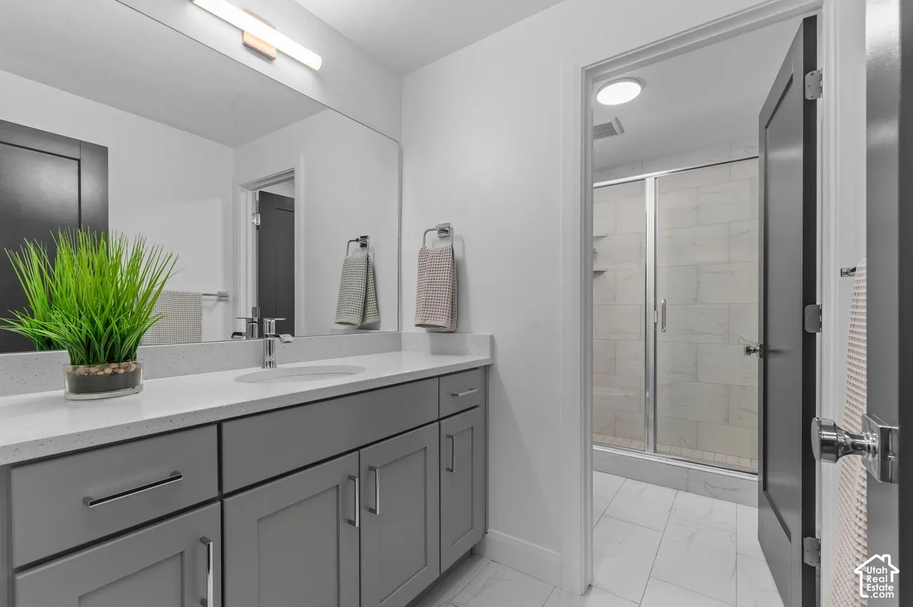 Grey shaker-style cabinetry with quartz countertop, undermount sink, full-width vanity mirror, tile flooring, and walk-in shower with tiled surround