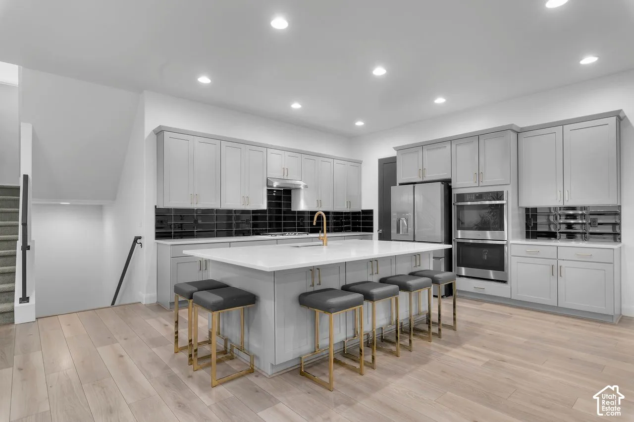 Kitchen featuring light grey shaker-style cabinetry, an expansive central island with stone countertop, black subway tile backsplash, stainless steel double wall oven, and wood-finish flooring
