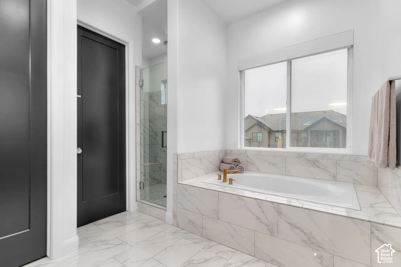 Deep soaking tub with a tiled surround, glass-enclosed walk-in shower with stone-patterned tile, matte black door hardware, and oversized tile flooring