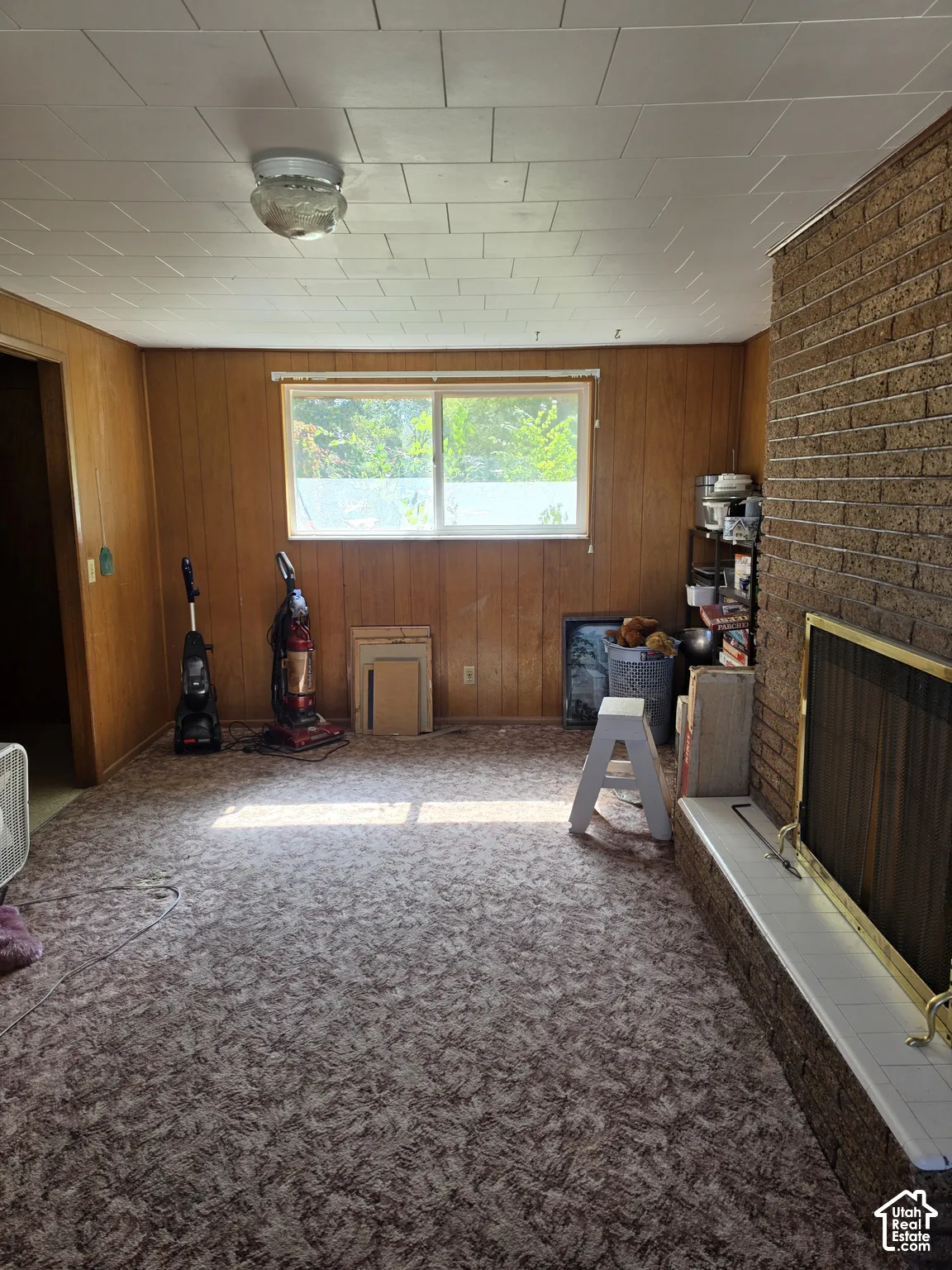 Full-height brick masonry fireplace with a tiled hearth and integrated screen, paneled wood wall finishes, textured carpeting, and a singular window with horizontal blind hardware