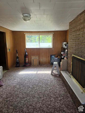 Full-height brick masonry fireplace with a tiled hearth and integrated screen, paneled wood wall finishes, textured carpeting, and a singular window with horizontal blind hardware