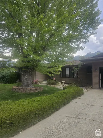 Exterior featuring horizontal siding, a paved driveway, mature tree with brick landscape edging, groomed shrubbery, and mountain backdrop