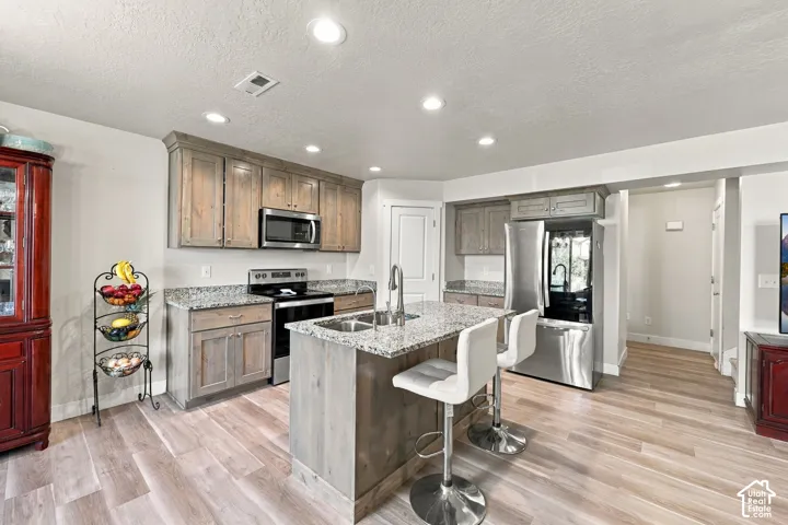 Kitchen featuring wood-finish cabinetry, stone-style countertops, stainless steel appliances, a center island with integrated sink, and wood-finish flooring