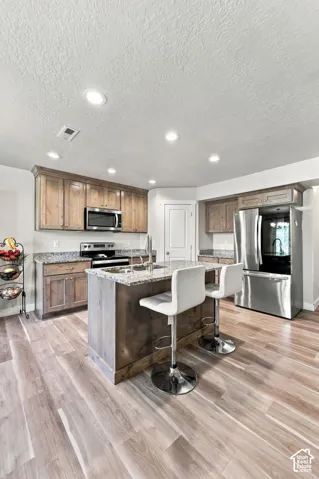 Kitchen featuring wood-finish cabinetry, stone-look countertops, central island with an integrated sink, stainless steel appliances, and wood-finish flooring