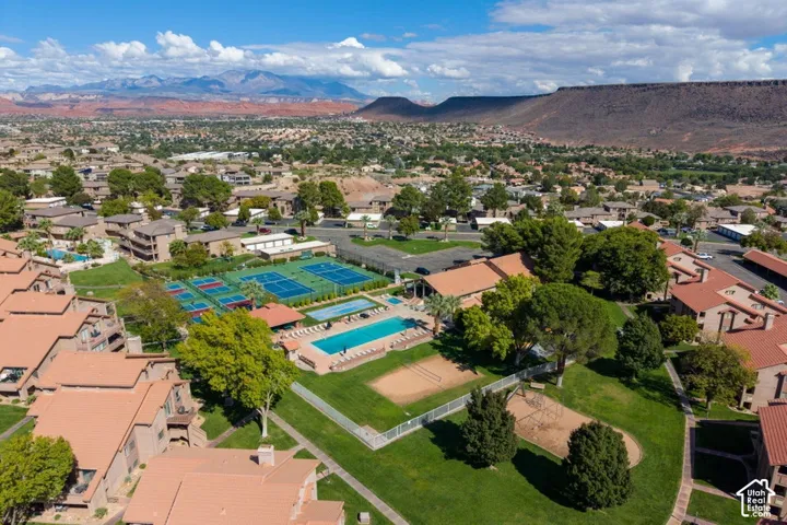 Aerial perspective of community grounds featuring a swimming pool, tennis courts, sand sports court, and expansive landscaped areas set against a mountain backdrop