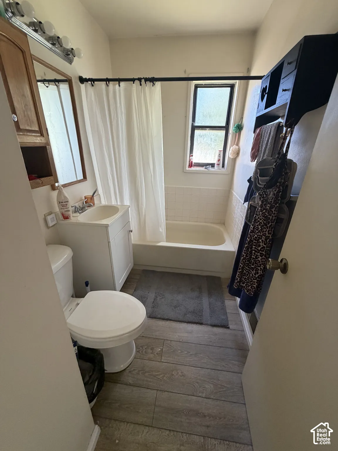 Bathroom featuring wood-finish flooring, a white pedestal vanity, a porcelain tub-and-shower combination with tiled surround, and a wall-mounted shelving unit