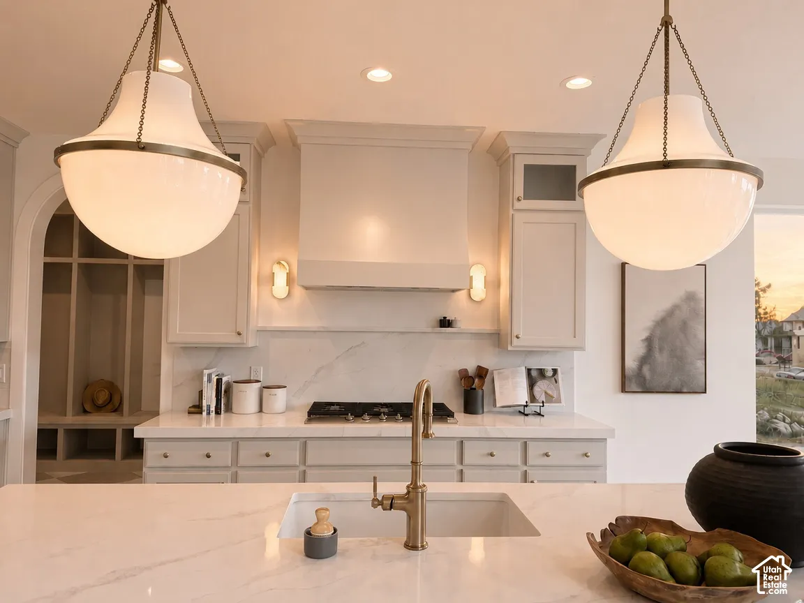 Kitchen featuring custom cabinetry, a stone-surfaced island with an integrated sink and metallic faucet, a matching stone backsplash, professional-style range hood, and dual oversized pendant light fixtures
