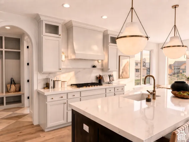 Custom kitchen featuring a large central island with light-toned solid surface countertops, ceiling-mounted pendant lighting, a built-in range hood, and cabinetry with integrated glass-paneled uppers