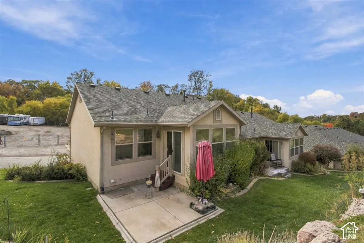 Rear exterior featuring a beige stucco facade, shingle roofing, a concrete patio, and landscaped grass grounds