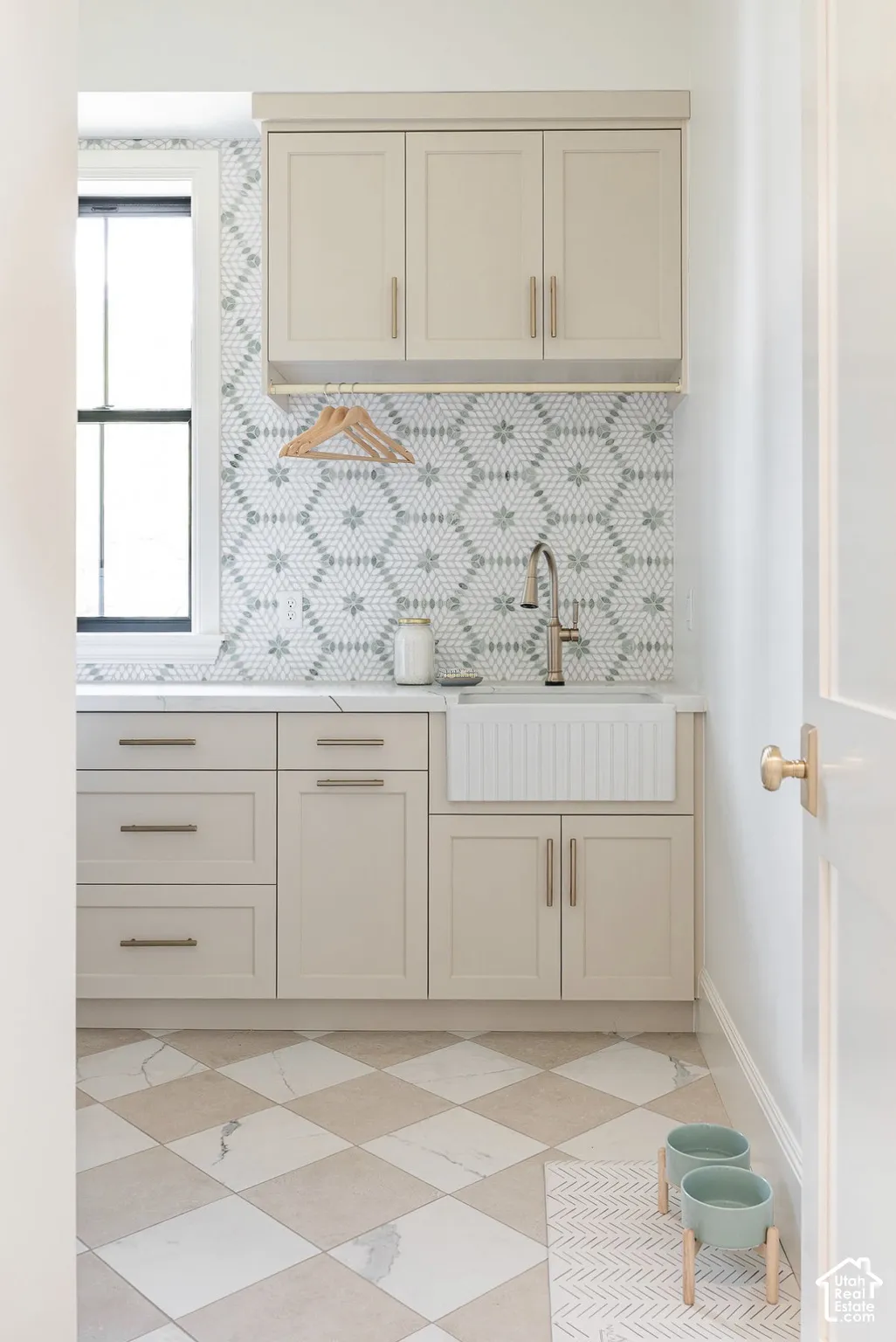 Custom cabinetry with metallic hardware, farmhouse-style apron-front sink, geometric patterned backsplash, and checkered stone-finish flooring