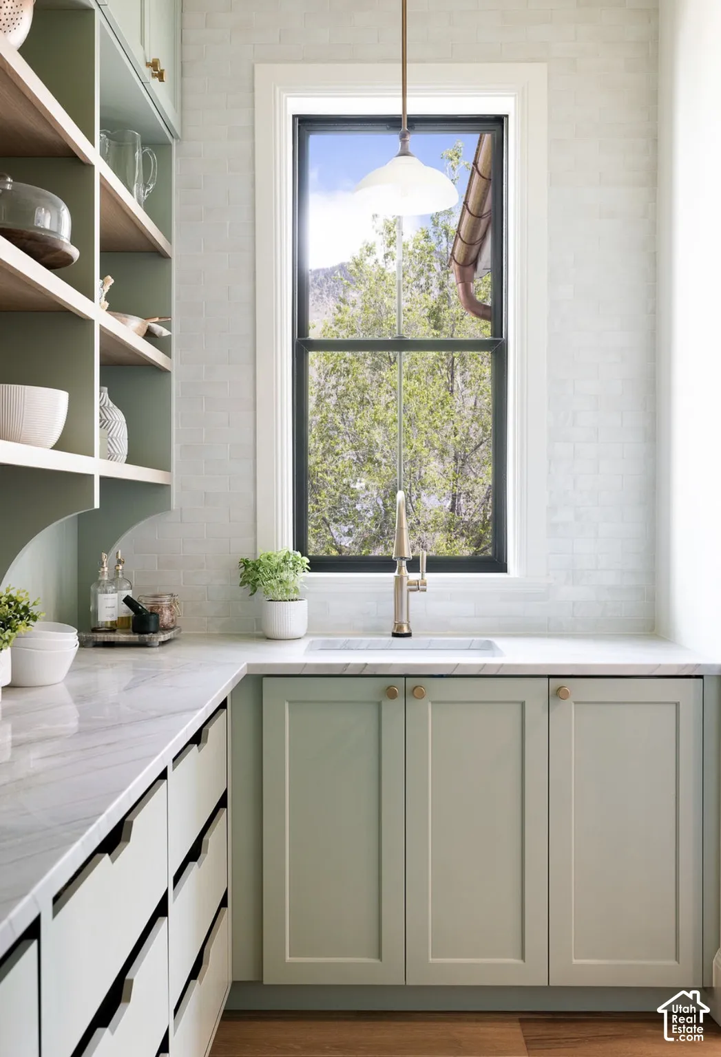 Custom light-green cabinetry, stone-finish countertops, undermount sink with a gooseneck faucet, white brick-pattern wall tile, and open wood shelving
