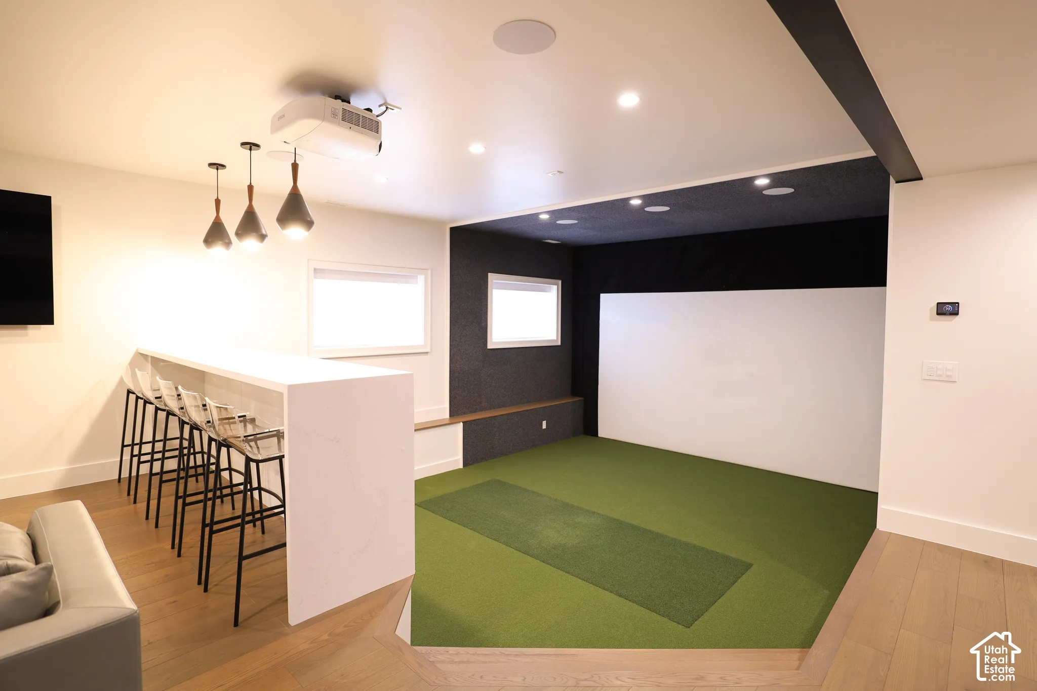 Custom interior featuring a synthetic turf floor with recessed putting strip, tiered bar with light-toned solid surface countertop, overhead pendant lighting, and ceiling-mounted projection system