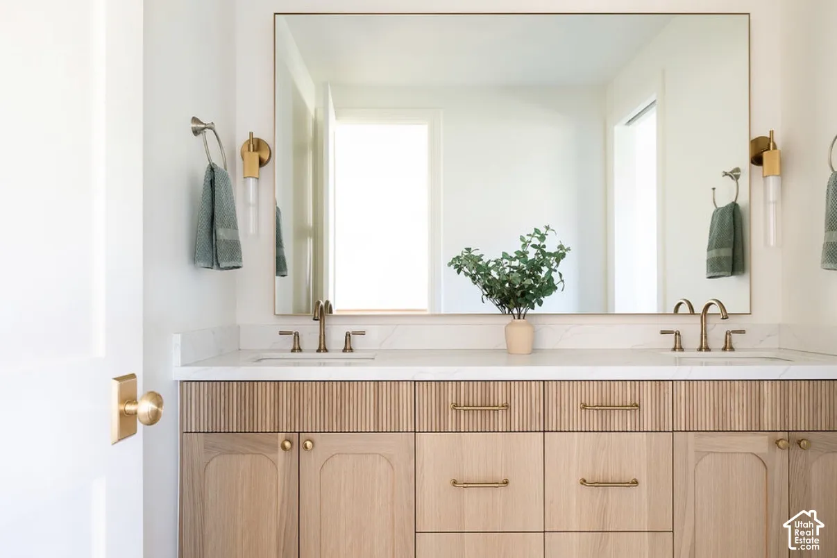 Dual-sink vanity featuring reeded cabinetry, stone-finish countertop, undermount sinks, brass-toned hardware, and rectangular wall mirror with integrated lighting