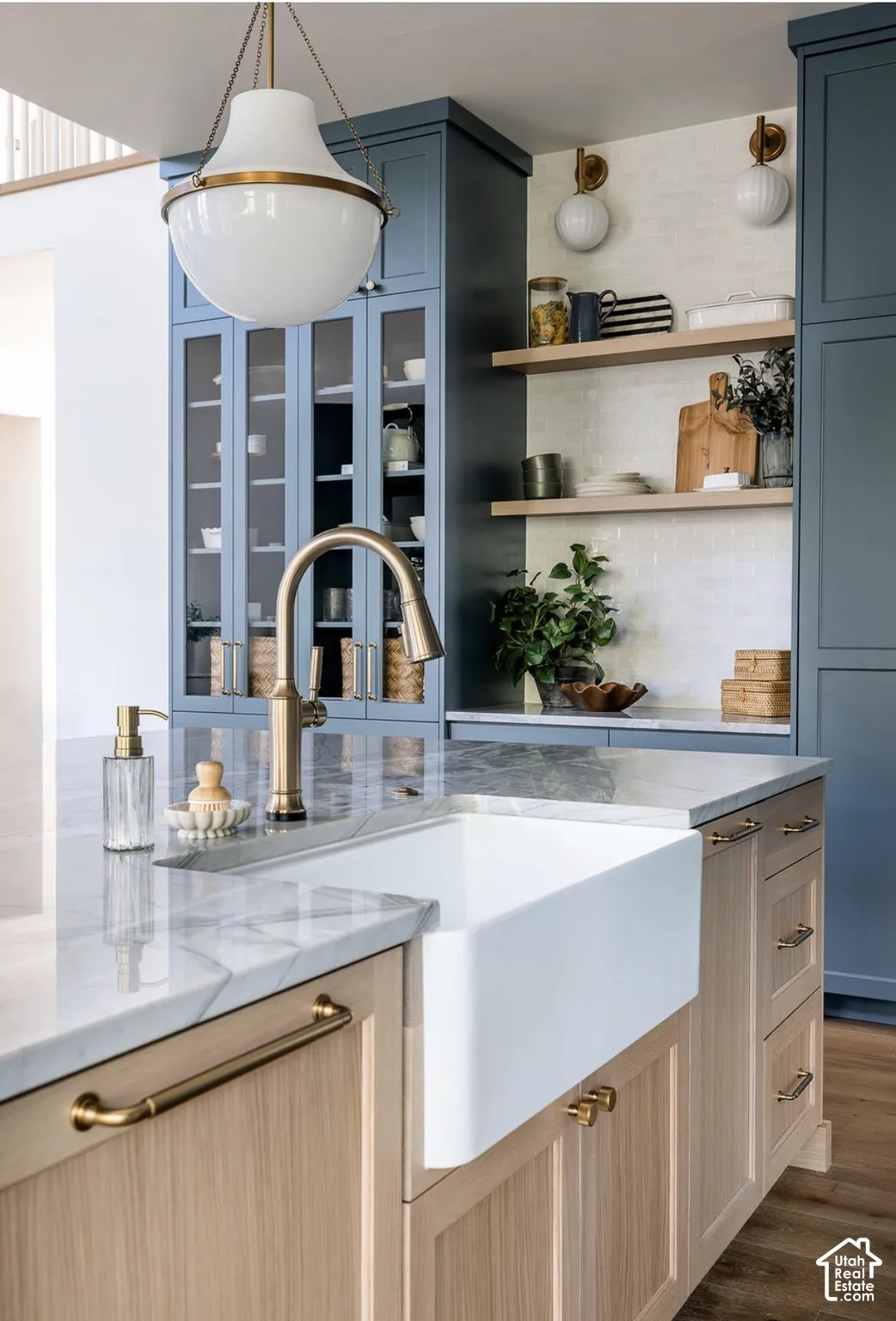 Kitchen island featuring a white farmhouse sink, stone countertop, wood-finish cabinetry, brass-toned hardware, and a pendant light fixture