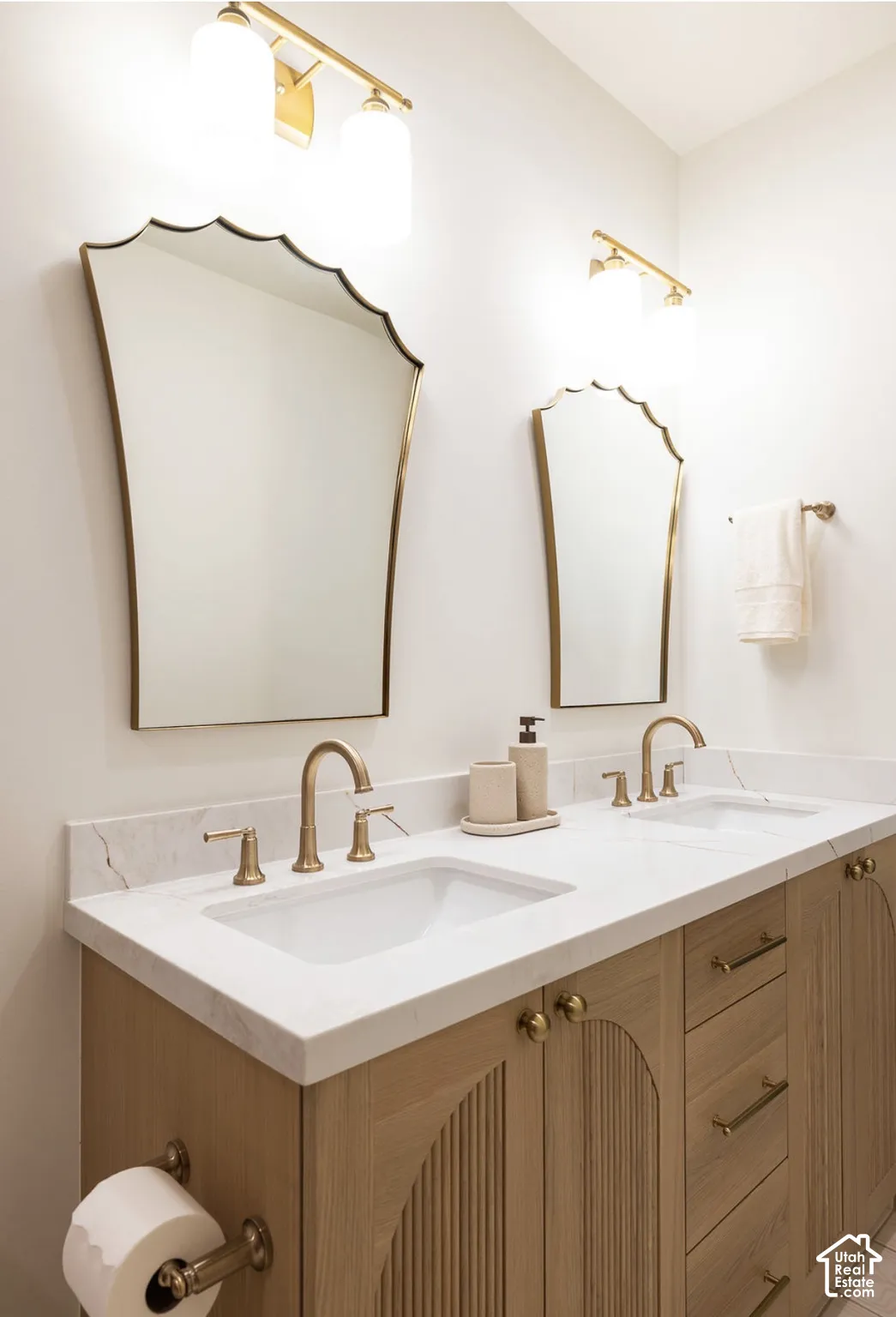 Double vanity featuring a white countertop with integrated sinks, wood-finish cabinetry with arched detailing, decorative wall mirrors, and brass-tone fixtures