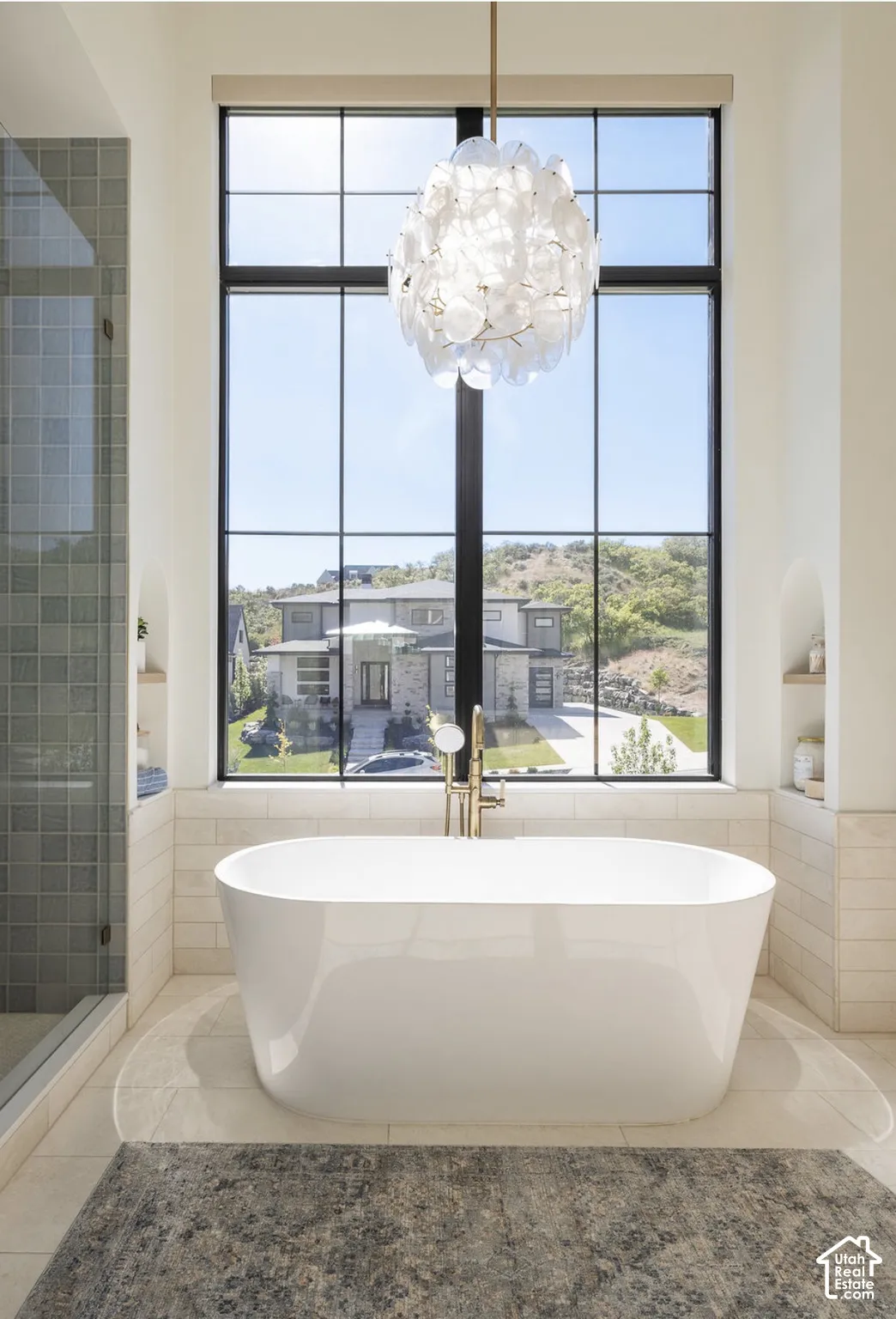 Freestanding soaking tub featuring a floor-mounted brass faucet, floor-to-ceiling multi-pane window, decorative chandelier, and built-in arched wall niches