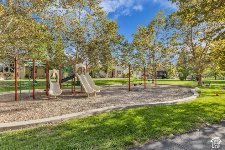 Recreational space featuring a playground structure with slides, a swing set, concrete curbing, and mature trees situated on a manicured lawn