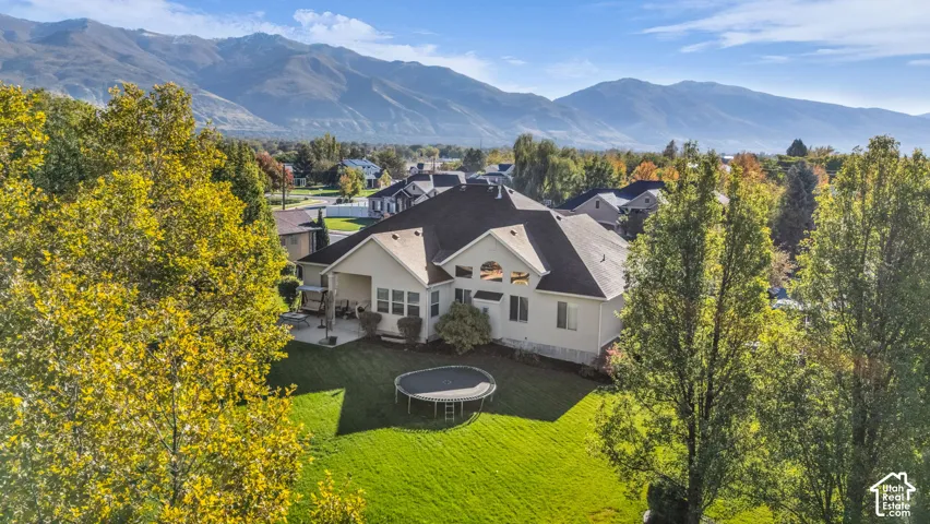 Rear exterior featuring a multi-gabled roofline, concrete patio, expansive lawn, mature perimeter trees, and a mountain backdrop