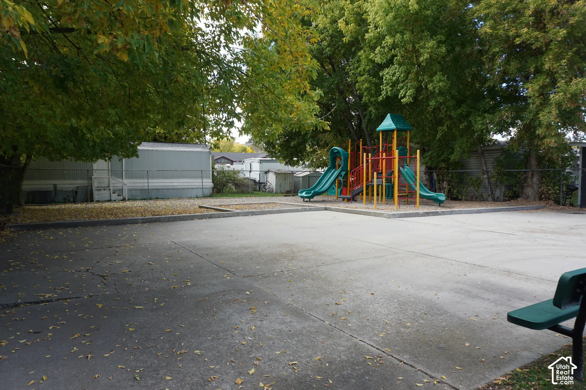 Outdoor recreation area featuring a concrete sports court, built-in playground equipment, chain-link perimeter fencing, and mature trees