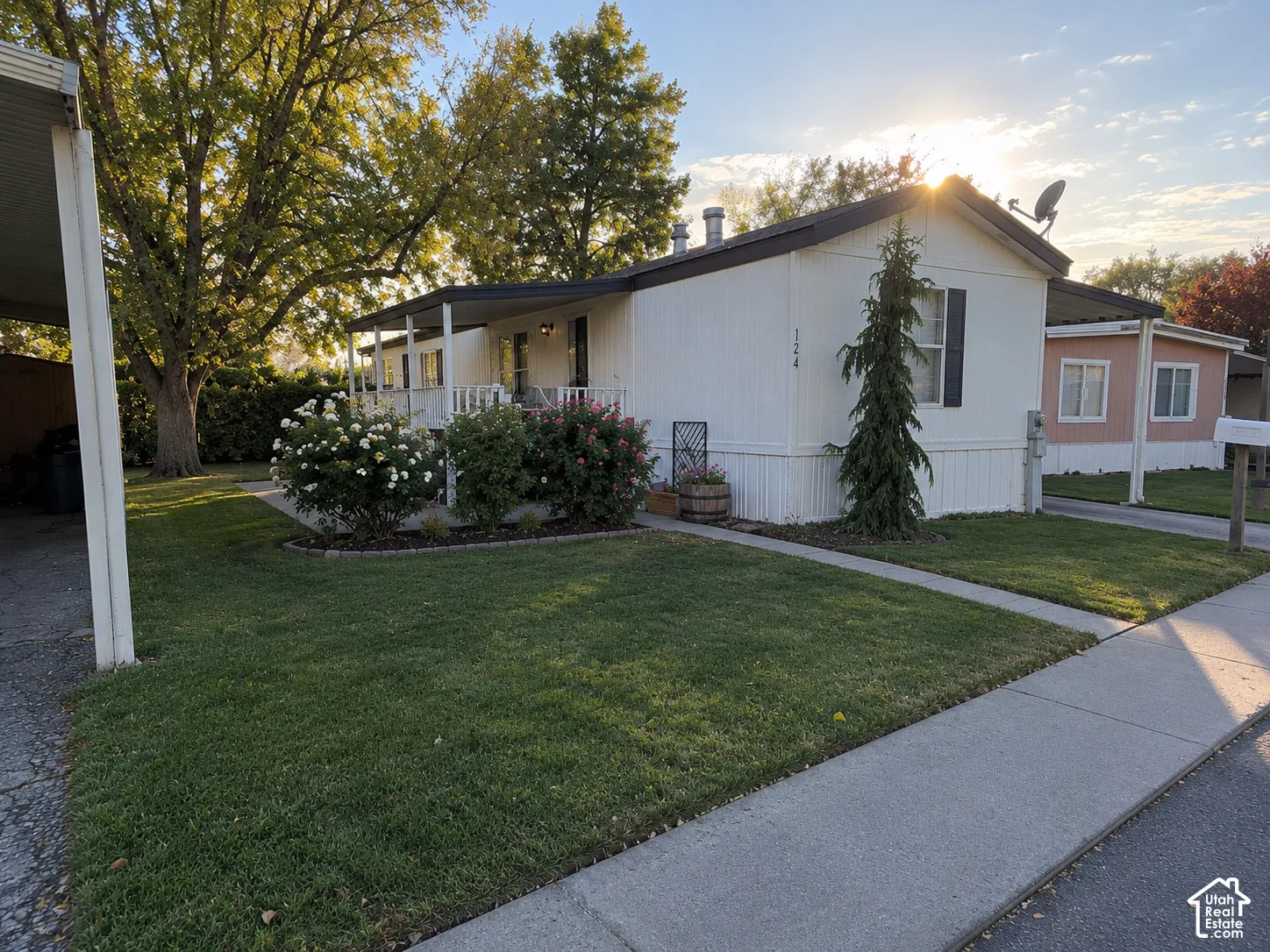 Exterior featuring white siding, a covered porch, mature trees, landscaped shrubbery, and a carport