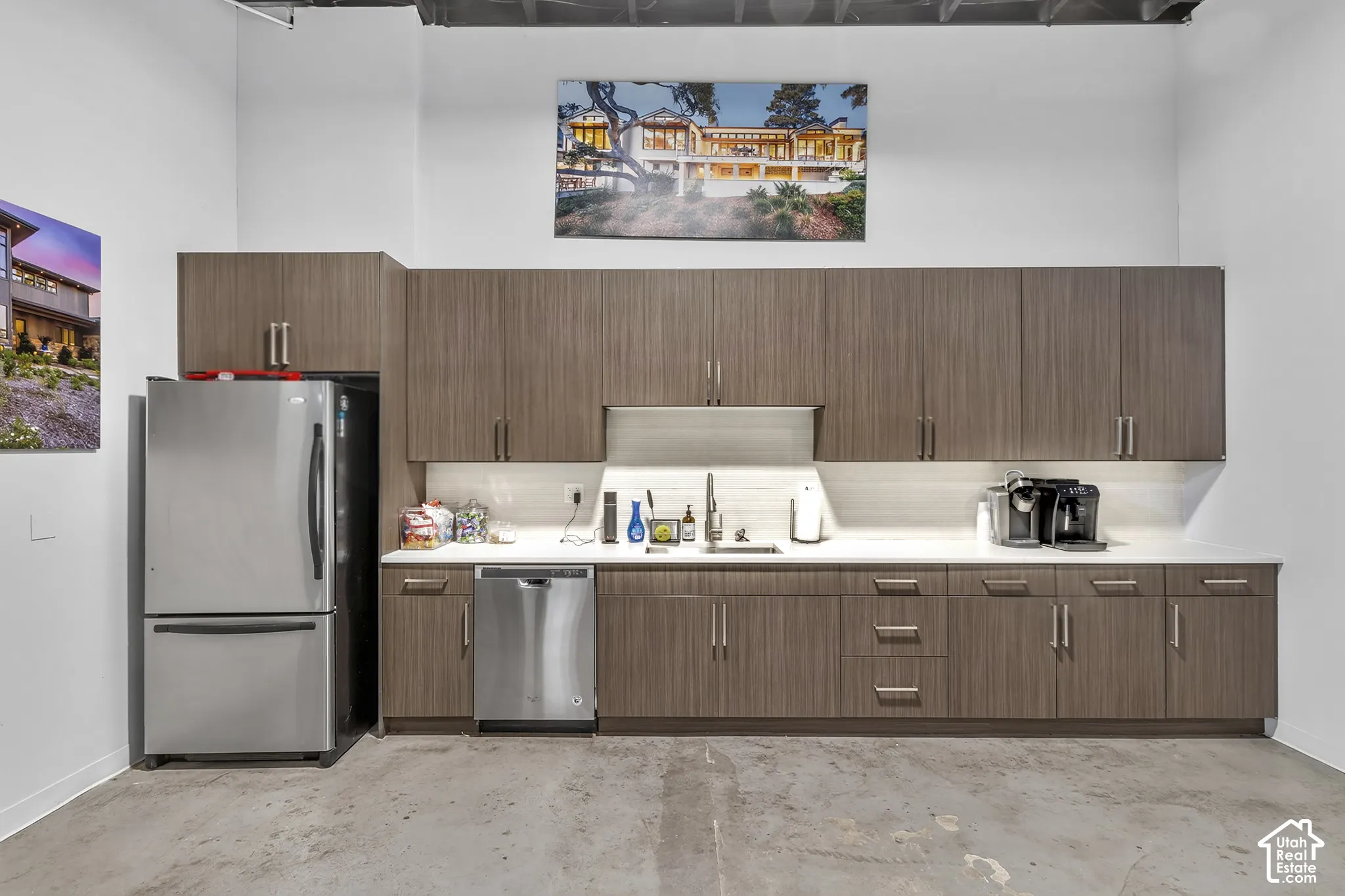 Wood-finish cabinetry featuring flat-panel doors and metallic hardware, light-toned stone-surface countertops, under-cabinet lighting, stainless steel dishwasher, and stainless steel bottom-freezer refrigerator