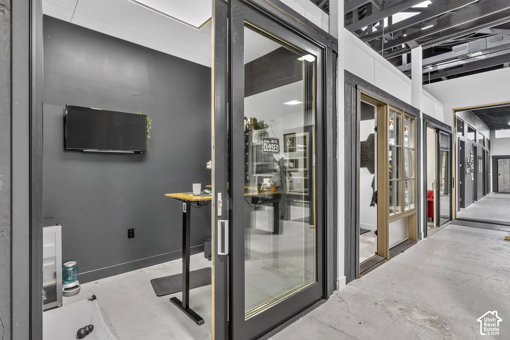 Enclosed private office space featuring glass-paned sliding doors, exposed ceiling beams, dark-toned interior walls, and recessed lighting