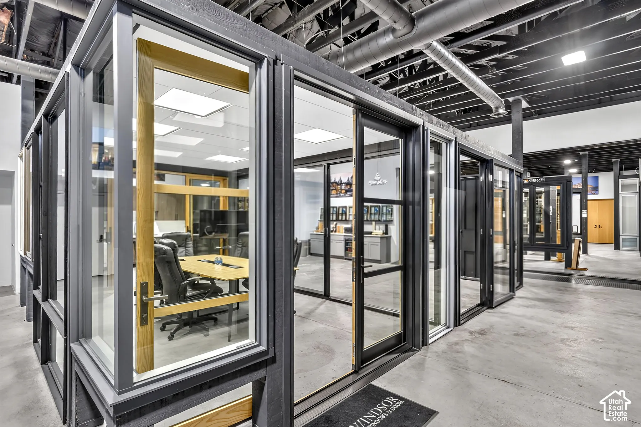 Enclosed glass office featuring dark-finished aluminum framing, interior wood trim, hinged glass door, and exposed industrial ceiling with overhead lighting