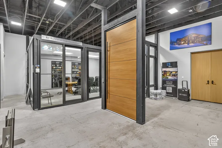 Oversized wood-finish pivot door with a vertical metal handle, industrial-style exposed ceiling, and floor-to-ceiling glass-enclosed partitioned space featuring black framing