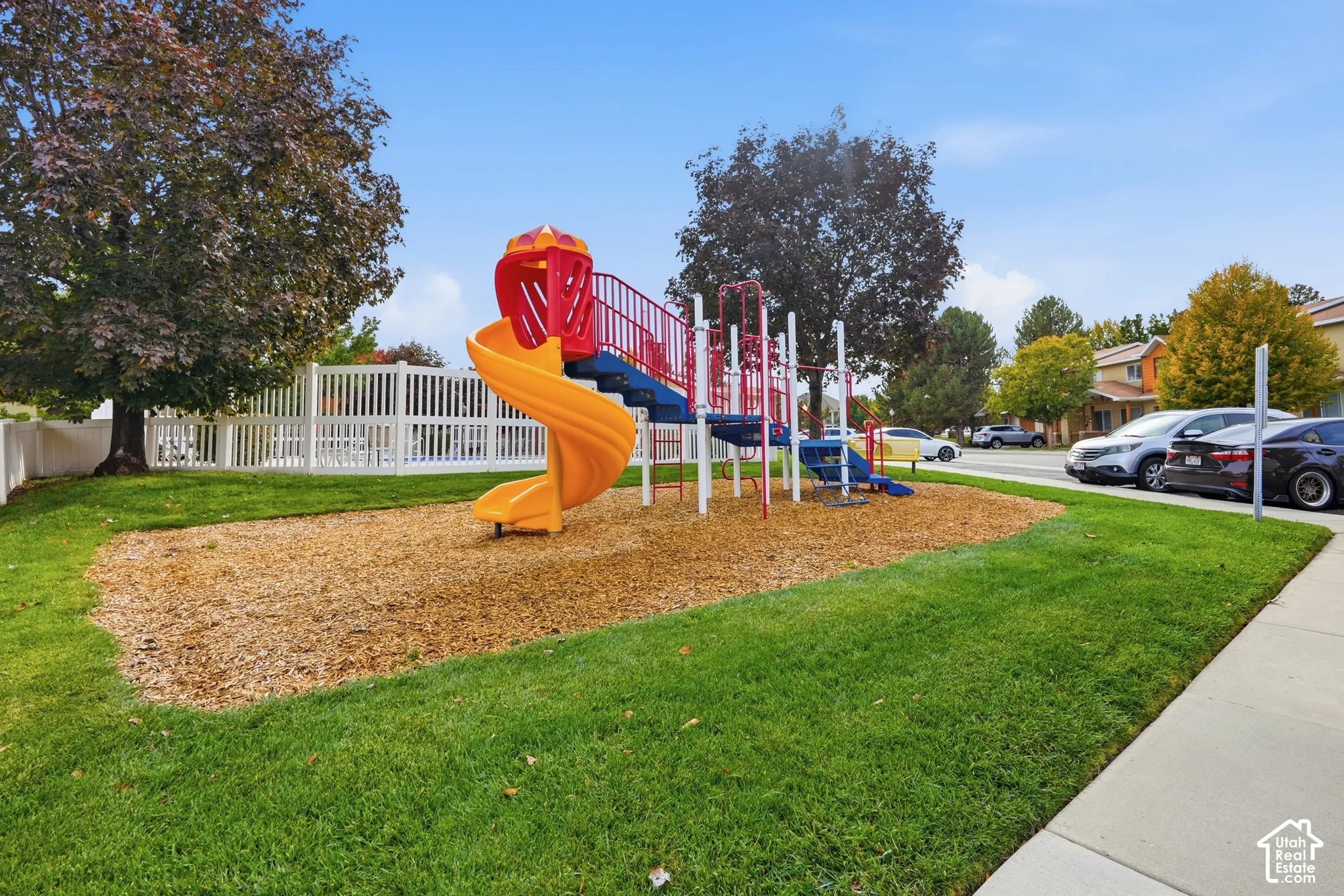 Recreational area featuring a playground structure with a spiral slide, wood chip ground covering, manicured lawn, and white vinyl privacy fencing