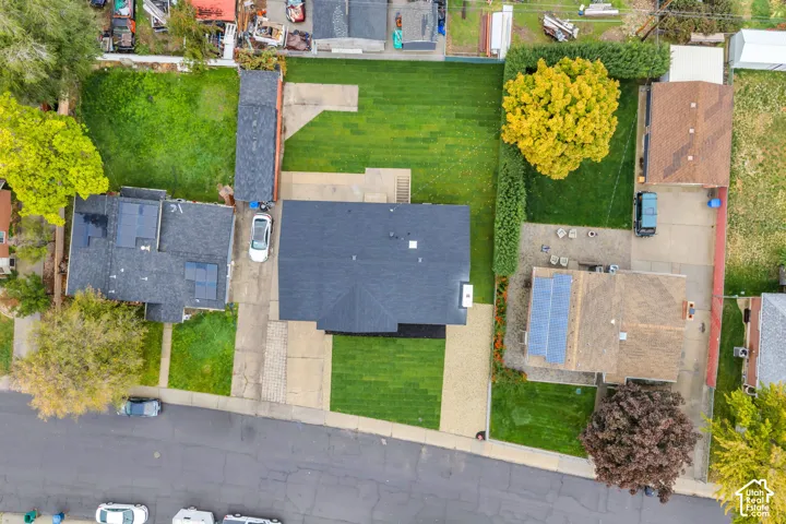 Aerial view featuring a residential property with a shingle roof, solar panel array, attached garage, paved driveway, and mature landscaping