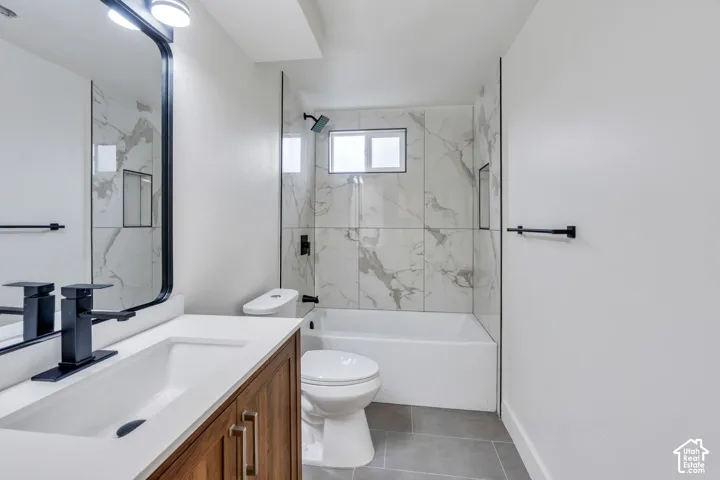Bathroom featuring a vanity with a white countertop, wood-finish cabinetry, matte black hardware, a tiled tub-shower surround with a stone-pattern finish, and grey floor tile