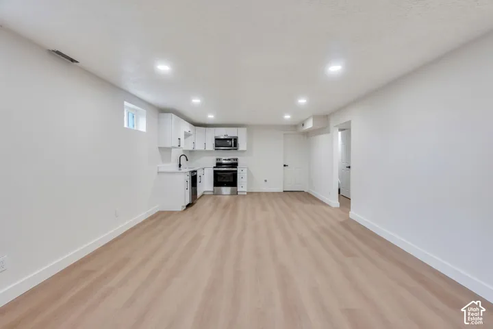 Open-concept area featuring wood-finish flooring, recessed lighting, and a kitchenette equipped with white cabinetry, stainless steel appliances, and a dark faucet