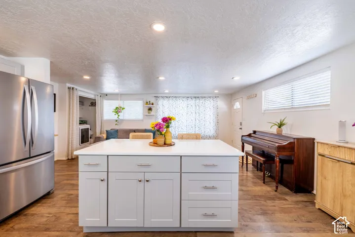 Kitchen island featuring a solid-surface countertop and soft-gray cabinetry, stainless steel refrigerator, recessed lighting, and wood-finish flooring