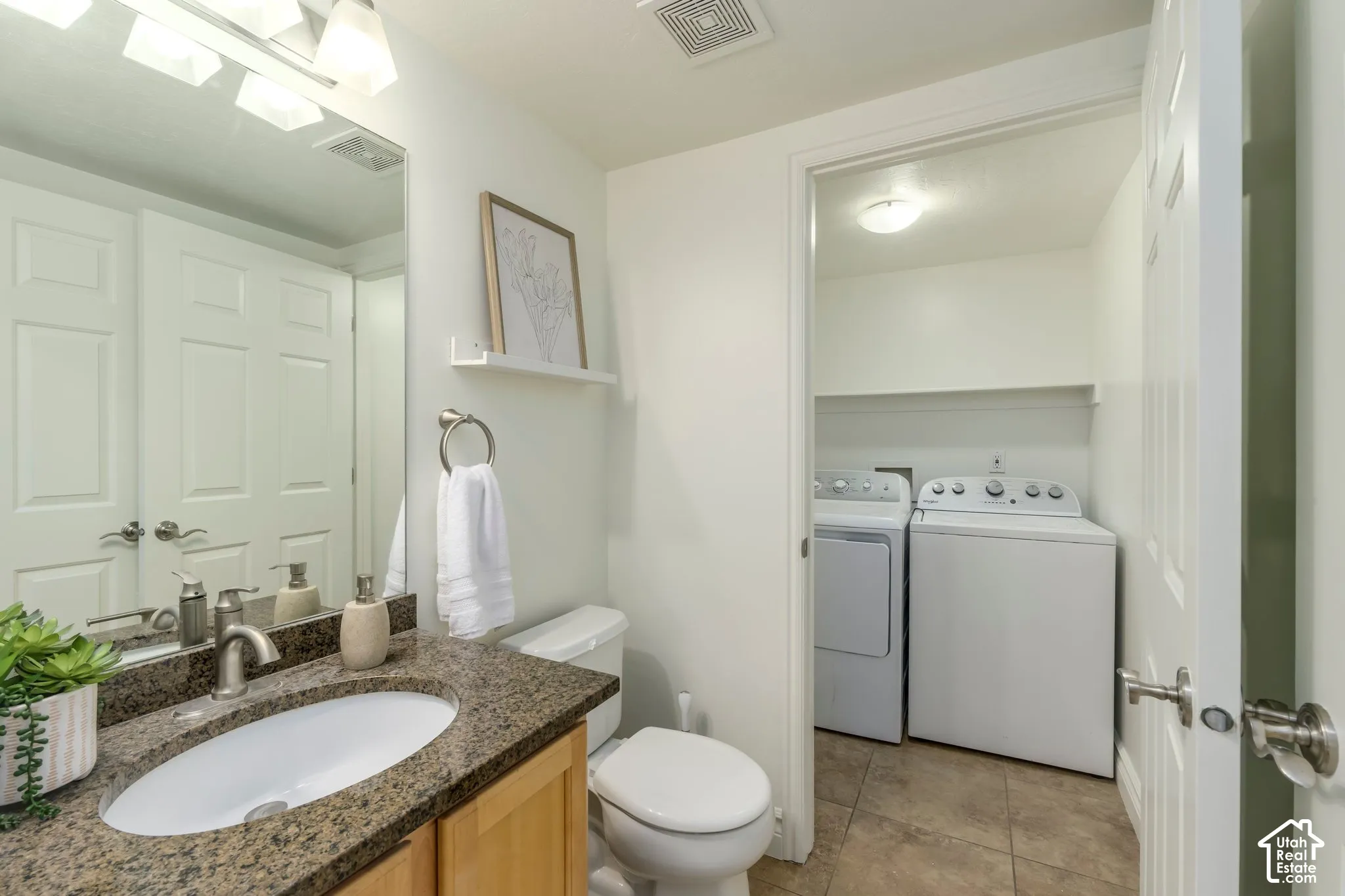 Bathroom featuring a stone-surface vanity with an integrated oval sink, full-width mirror, and three-bulb vanity light fixture, plus an adjoining utility area with tile flooring and built-in shelving