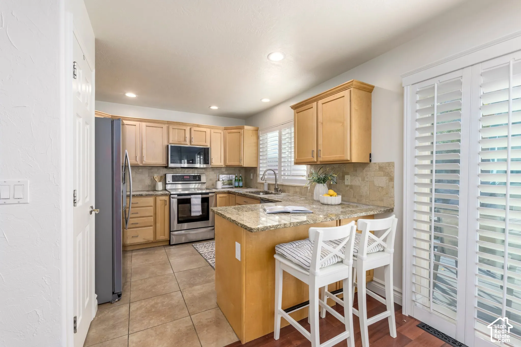Kitchen featuring light wood cabinetry, stone countertops, stainless steel appliances, tile backsplash, and plantation shutters