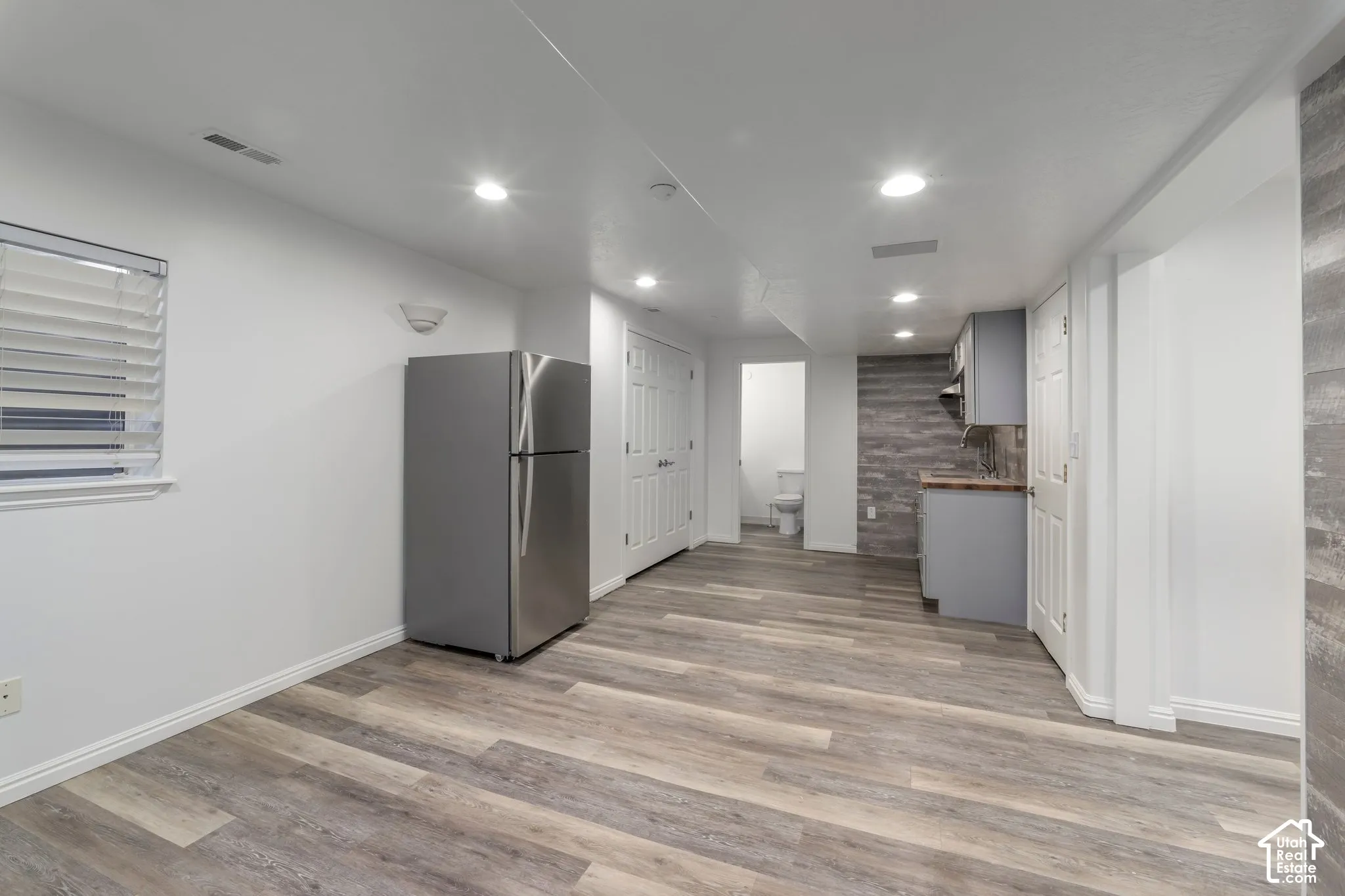 Wood-finish flooring, stainless steel refrigerator, kitchenette with grey cabinetry, wood-textured accent wall, and recessed lighting
