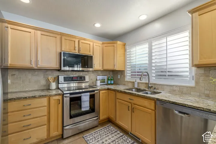 Kitchen featuring natural-finish wood cabinetry, stone countertops, tiled backsplash, stainless steel appliances, and plantation shutters