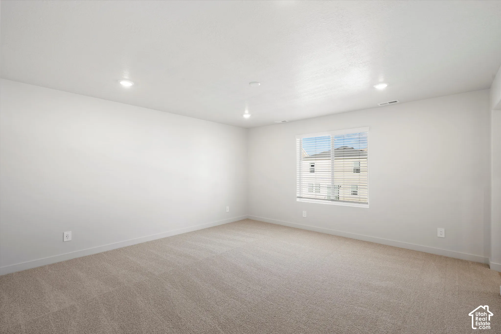 Neutral-toned interior space featuring wall-to-wall carpeting, recessed ceiling lighting, and a single-pane window with integrated blinds