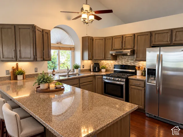 Kitchen featuring warm-toned cabinetry, an expansive granite surface island, stainless steel appliances, a multi-textured backsplash, and an arched window. Glazed cabinets are lighter in person.