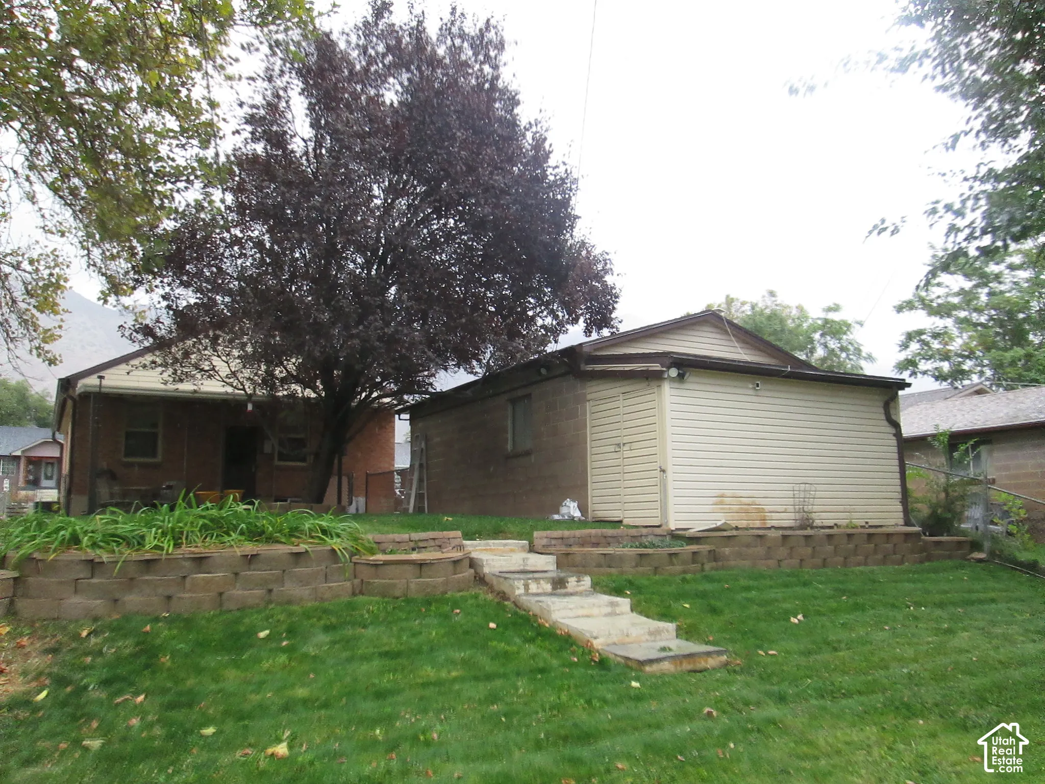 Rear exterior featuring a brick and vinyl-sided structure, tiered concrete block retaining walls, stone stairs, and mature trees