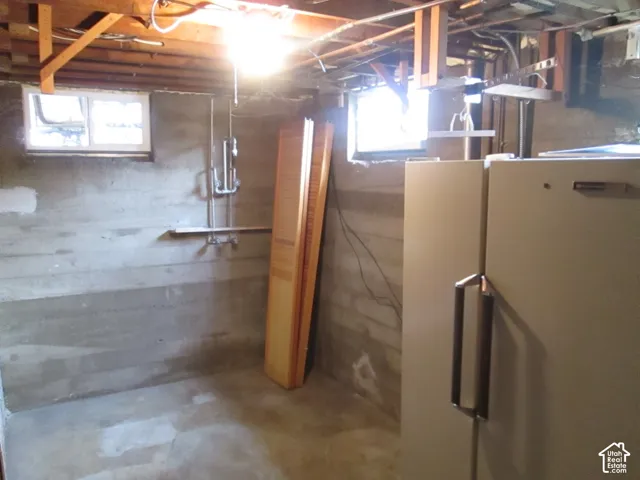 Lower-level utility area featuring exposed wood joist ceilings, concrete wall construction, dual sliding windows, and a wall-mounted plumbing fixture