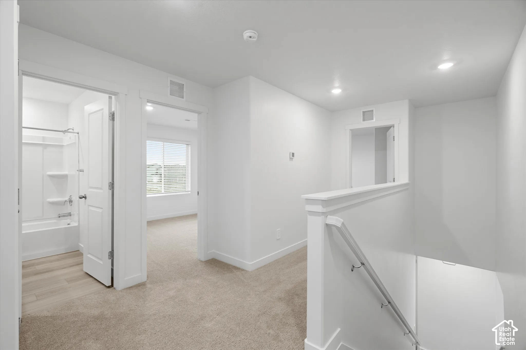 Neutral interior hallway featuring plush carpeting, recessed ceiling lighting, a white painted railing with a metal handrail, and entryways to a carpeted room and a full bathroom with a tub-shower combination
