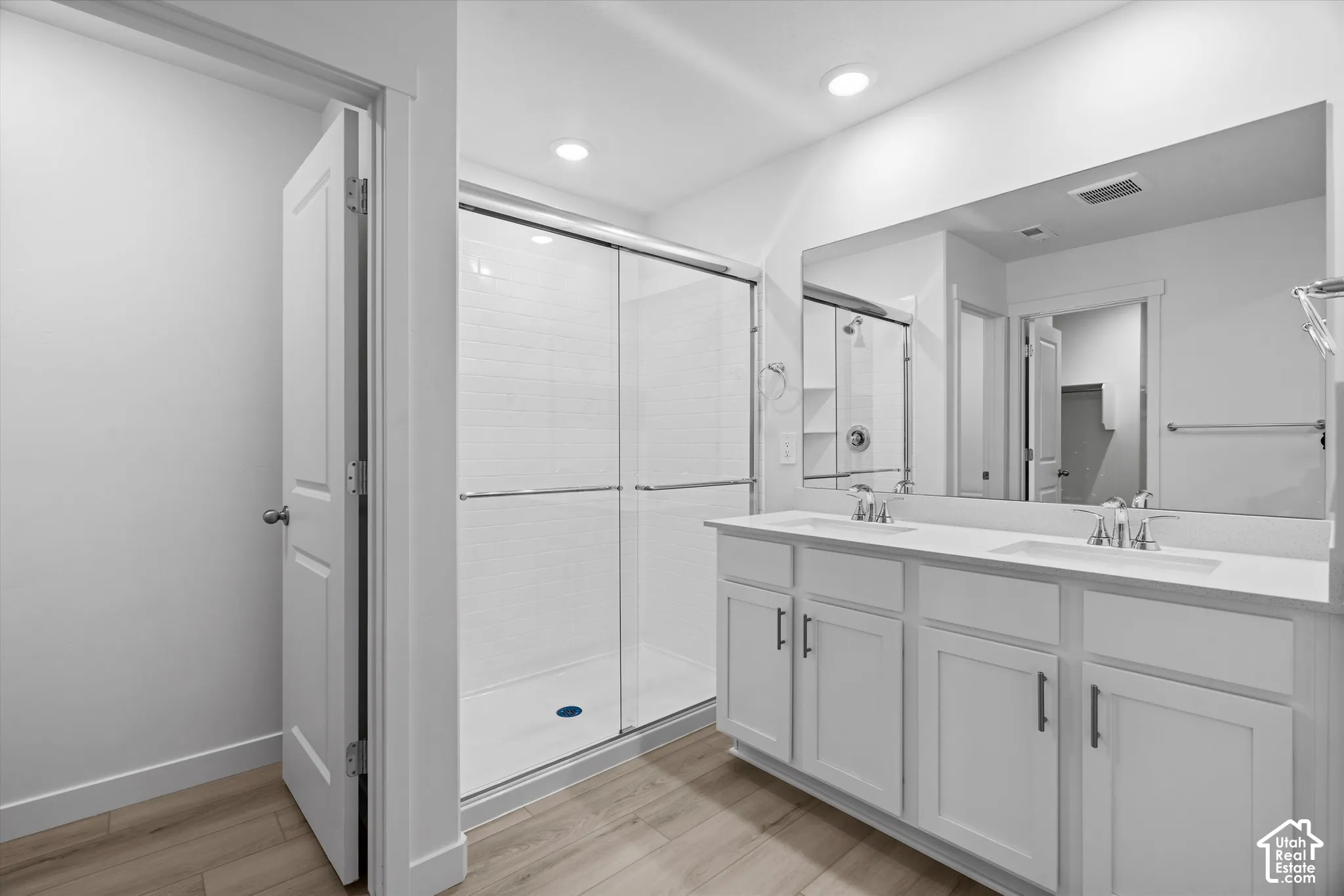Dual-sink vanity with white cabinetry and countertop, large wall-mounted mirror, and glass-enclosed shower with white tiled surround and wood-finish flooring