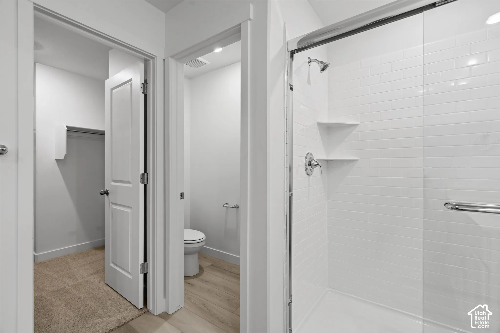 Walk-in shower with white subway tile surround, frameless glass door, and built-in shelving, complemented by a water closet and reach-in storage area with carpeted flooring