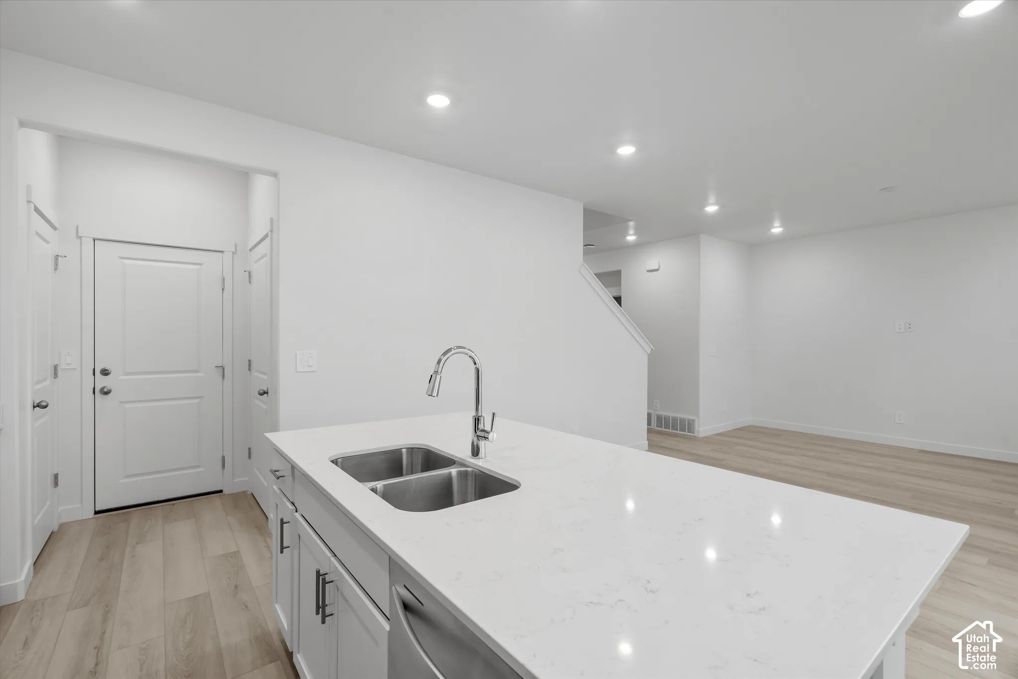 Kitchen island featuring a quartz countertop, under-mount dual-basin stainless steel sink, gooseneck faucet, and white shaker-style cabinetry, complemented by wood-finish flooring and recessed lighting