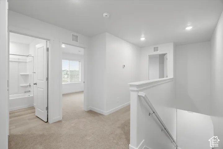 Neutral interior hallway featuring plush carpeting, recessed ceiling lighting, a white painted railing with a metal handrail, and entryways to a carpeted room and a full bathroom with a tub-shower combination